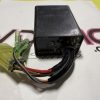 Black rectangular electrical part with multiple colored wires and a yellow connector, resting on a white surface with pink letters in the background.