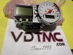 Disassembled car tachometer gauge cluster with orange needle, two digital displays, and exposed circuit board on a pink bubble wrap background.