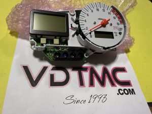 Disassembled car tachometer gauge cluster with orange needle, two digital displays, and exposed circuit board on a pink bubble wrap background.