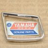 Chrome rectangular frame badge with a Yamaha Genuine Parts sticker showing the Yamaha logo and text 'Genuine Parts'.