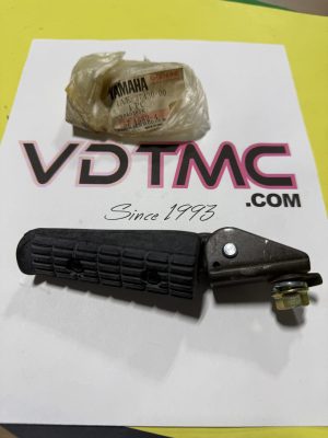 Black motorcycle footpeg with mounting bracket lying on a white sheet; a small Yamaha-branded bag sits nearby.