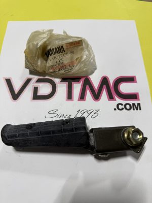 Small crumpled plastic bag with a Yamaha Genuine Parts label sits on a white surface with a large pink-and-black logo in the background; a black motorcycle foot peg is in the foreground.
