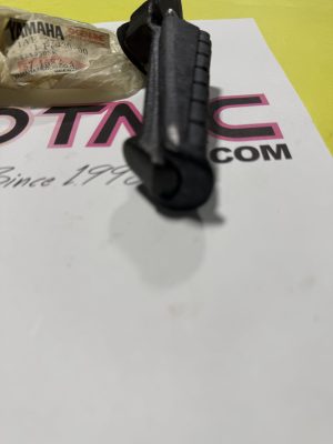 Close-up of a black plastic component resting on a white poster with pink lettering, with a crumpled Yamaha Genuine bag on the left and a yellow background.