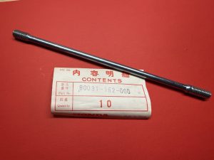 Long metal bolt with threaded ends lying diagonally on a red surface, partly covering a white contents label.