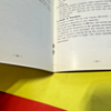 s-l140-2 Close-up of an open book with text on white pages, resting on a blue surface with yellow and red areas nearby.