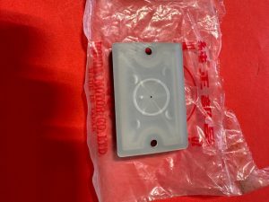 Grey plastic wall plate with a circular dial recess and two screw holes, resting on a clear plastic bag on a red surface