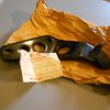 Black automotive bracket with multiple circular holes, lying on crumpled brown paper, with a Honda contents tag attached/nearby.
