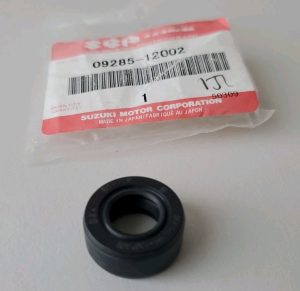 Black rubber oil seal with a hollow center placed on a light surface next to a Suzuki Motor Corporation parts bag with a barcode and red label