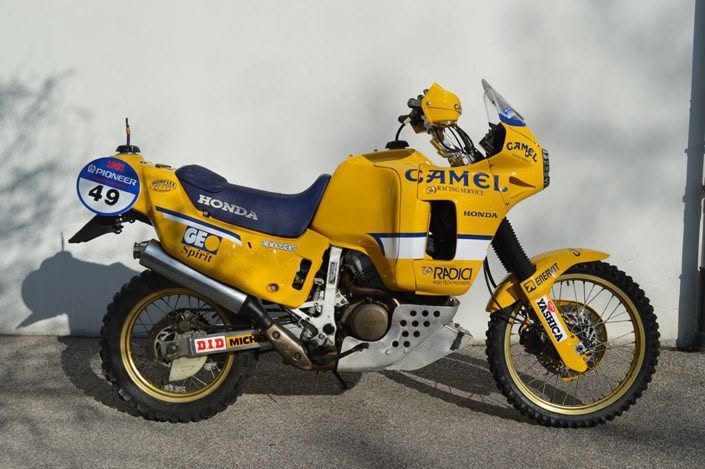Yellow Honda off-road racing motorcycle with Camel sponsorship, number 49, parked on a paved surface with a white wall in the background.