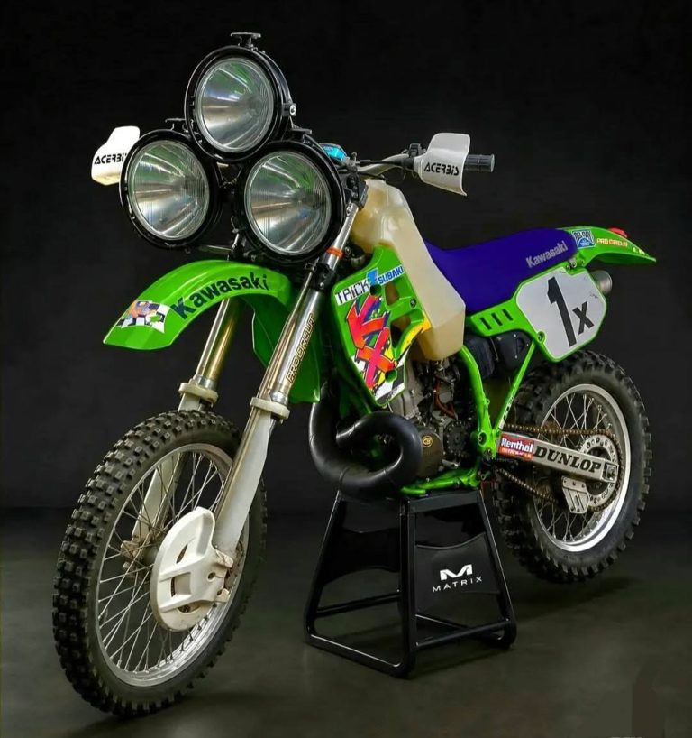 Kawasaki dirt/trial bike in green with blue seat, mounted on a stand against a black background, showing racing decals and large front lights.
