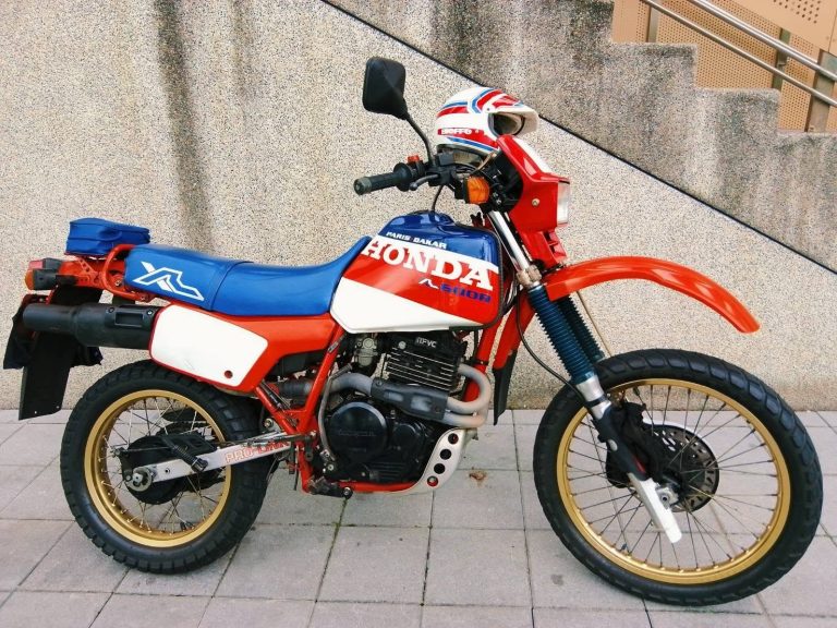 Red and blue Honda Paris-Dakar dirt bike with gold wheels parked on a sidewalk; helmet rests on the handlebars.