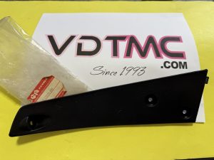Black plastic interior car trim piece with a mounting hole, resting on a white brochure on a yellow surface, with a red sticker nearby.