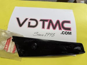 Black plastic automotive part on a bright yellow surface with a red-and-white Suzuki label and a white sheet beside it.