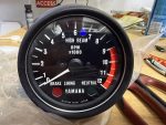 Close-up of a motorcycle tachometer face showing RPM x1000, numbers 0–12, with Yamaha branding and high-beam indicator on a workshop bench.