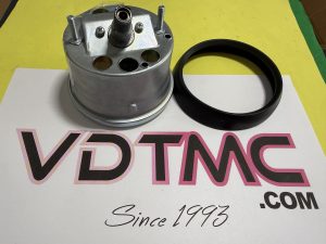 Metal cylindrical mechanical part with a central threaded shaft and several holes, placed next to a black circular ring on a yellow surface with printed text in the background.