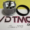Metal cylindrical mechanical part with a central threaded shaft and several holes, placed next to a black circular ring on a yellow surface with printed text in the background.