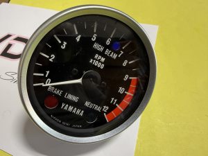 Close-up of a motorcycle tachometer displaying RPM x1000, with numbers 0–12, a blue indicator, redline area, and Yamaha/Nippon Seiki branding visible.