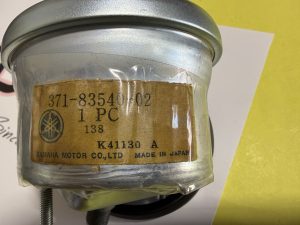 Close-up of a gray metal can/part with a worn brown label taped on; label shows parts text and Yamaha branding partially visible (Made in Japan).