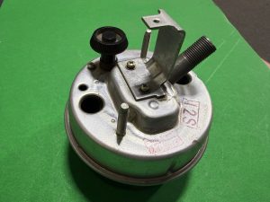 Close-up of a metal mechanical component with a knurled black adjustment knob, threaded rod, and mounting bracket on a circular base against a green background.