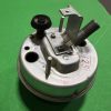 Close-up of a metal mechanical component with a knurled black adjustment knob, threaded rod, and mounting bracket on a circular base against a green background.