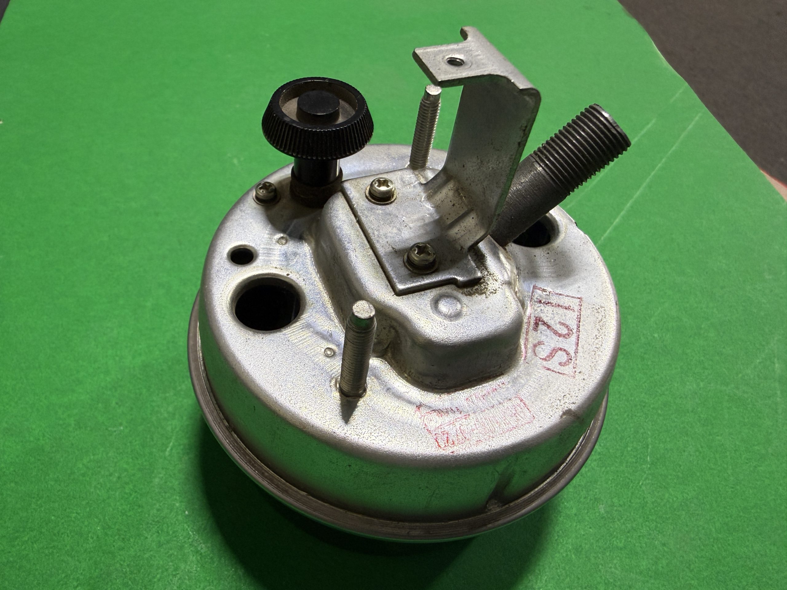 Close-up of a metal mechanical component with a knurled black adjustment knob, threaded rod, and mounting bracket on a circular base against a green background.