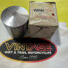 Metal motorcycle piston beside a worn Yamaha parts box on a yellow surface.