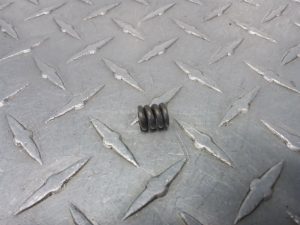 Small black helical compression spring resting on a gray diamond-plate metal surface.
