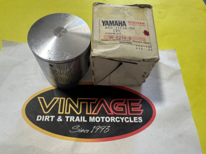 Metal motorcycle piston beside a worn Yamaha parts box on a yellow surface.