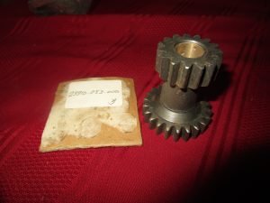 Two dark metal spur gears stacked on a short shaft, with a red fabric background and a small worn tag nearby.