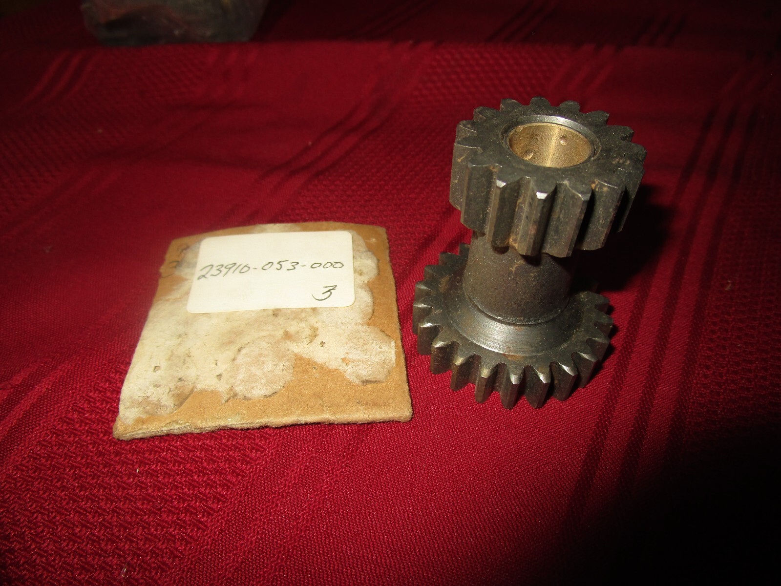 Two dark metal spur gears stacked on a short shaft, with a red fabric background and a small worn tag nearby.