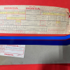 Honda parts label with stamped form and red box artwork, placed on a red surface, and a blue plastic item resting on a metal box below.