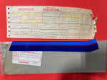 Honda parts label with stamped form and red box artwork, placed on a red surface, and a blue plastic item resting on a metal box below.