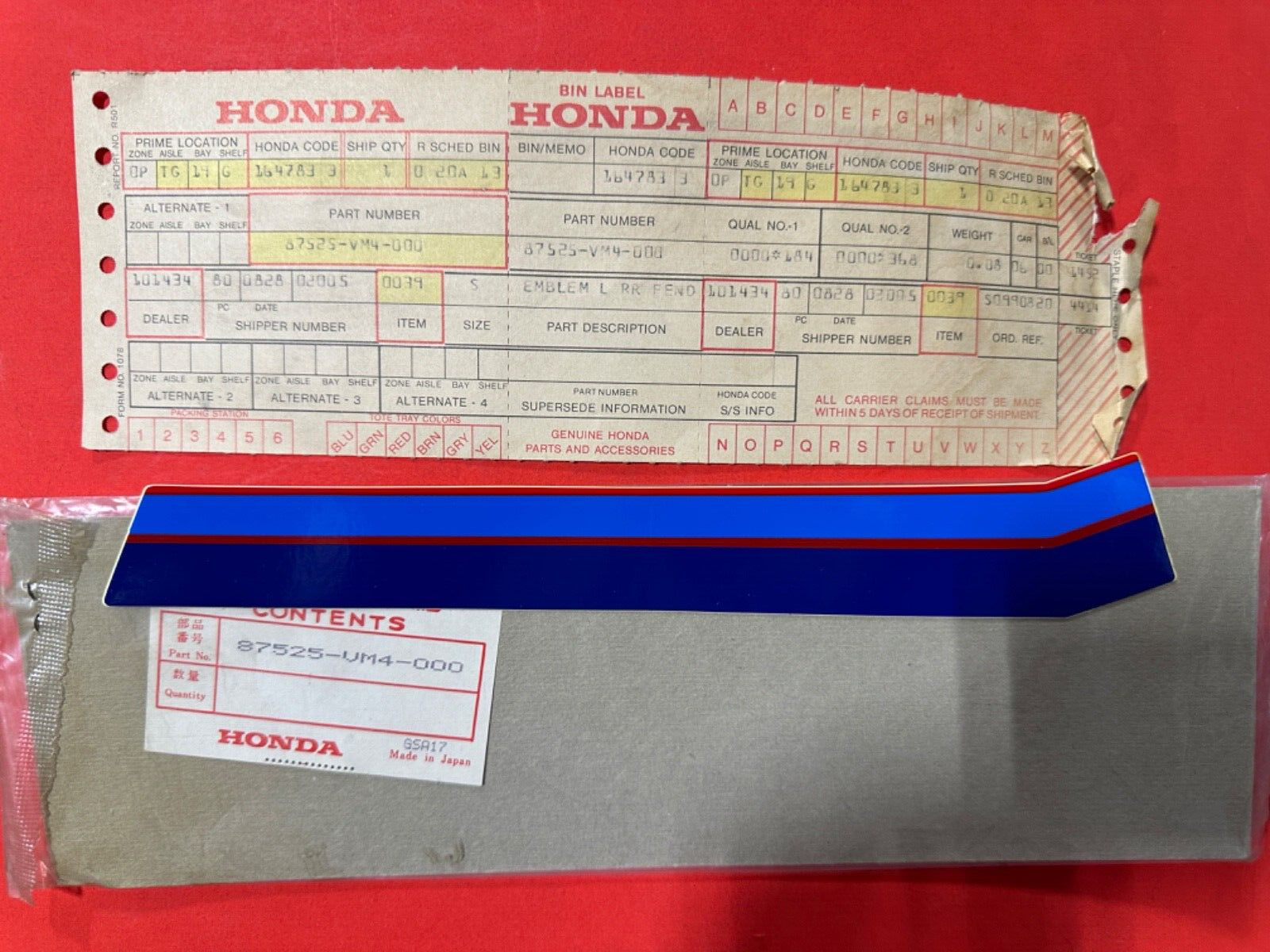 Honda parts label with stamped form and red box artwork, placed on a red surface, and a blue plastic item resting on a metal box below.