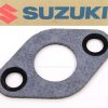 Suzuki logo on cardboard above a gray gasket with two small holes and one large central opening, likely an engine gasket for a Suzuki vehicle.