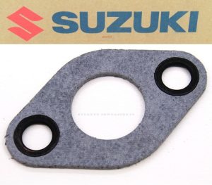 Suzuki logo on cardboard above a gray gasket with two small holes and one large central opening, likely an engine gasket for a Suzuki vehicle.