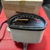 Analog automotive gauge with a curved scale and yellow markings, needle near 0, housed in light gray casing on a red surface with a cardboard box in the background.