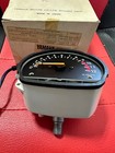 Analog automotive gauge with a curved scale and yellow markings, needle near 0, housed in light gray casing on a red surface with a cardboard box in the background.