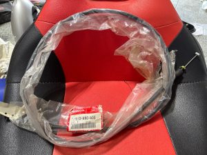 Close-up of a red and black motorcycle seat with a clear plastic bag and a loose cable resting on it, Honda tag visible.