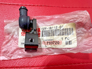 Small black plastic automotive component with a side nozzle and mounting hole on a pink textured surface, resting on a bag with a barcode label.
